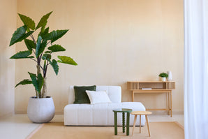 Ein minimalistisches Wohnzimmer mit der künstlichen Calocasia-Pflanze 210 cm, einem weiß strukturierten Sofa mit grünen und weißen Kissen, zwei kleinen Beistelltischen, einem beigen Teppich und einem hölzernen Konsolentisch vor einer hellbeigen Wand.