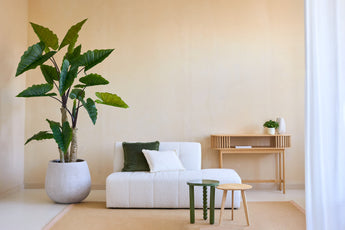 Ein minimalistisches Wohnzimmer mit der künstlichen Calocasia-Pflanze 210 cm, einem weiß strukturierten Sofa mit grünen und weißen Kissen, zwei kleinen Beistelltischen, einem beigen Teppich und einem hölzernen Konsolentisch vor einer hellbeigen Wand.