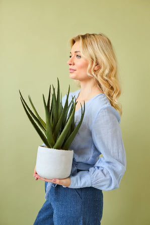 Eine Frau mit langen blonden Haaren in hellblauer Bluse und blauer Hose hält die Kunstpflanze Aloe Vera 43 cm in einem Topf vor einem schlichten hellgrünen Hintergrund.