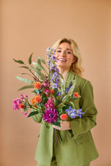 Eine lächelnde blonde Person in einem grünen Anzug hält den farbenfrohen künstlichen Seidenblumenstrauß von Svenski vor einem schlichten, hellbraunen Hintergrund.