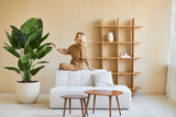 Eine Frau in einem braunen Overall sitzt auf der Rückenlehne eines weißen Sofas und berührt das Blatt einer künstlichen Strelitzia Real Touch Deluxe 180 cm Pflanze in einem minimalistischen Wohnzimmer mit Holztischen und Regalen.