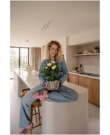Eine Frau mit lockigen blonden Haaren, Jeans und gestreiften Socken, sitzt lächelnd auf der Küchentheke und hält die künstliche Gardenie UV 39 cm – eine Topfpflanze mit gelben Blüten. Im Hintergrund sieht man eine moderne Küche mit Holzschränken und Regalen.