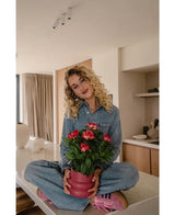 Eine Frau mit lockigen blonden Haaren, trägt einen Jeansoverall und rosa Turnschuhe, sitzt mit gekreuzten Beinen auf einer Küchentheke und lächelt, während sie die künstliche Gardenie UV 39 cm in Fuchsia hält.