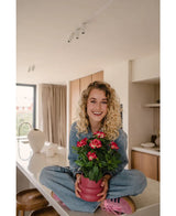 Eine lächelnde Frau mit lockigen blonden Haaren sitzt im Schneidersitz auf einer hellen, modernen Arbeitsplatte und hält die künstliche Gardenia UV 39 cm Fuchsia – eine rosa Topfpflanze mit leuchtend roten Blüten. Sie trägt Jeans, ein blaues Hemd und rosa Schuhe.