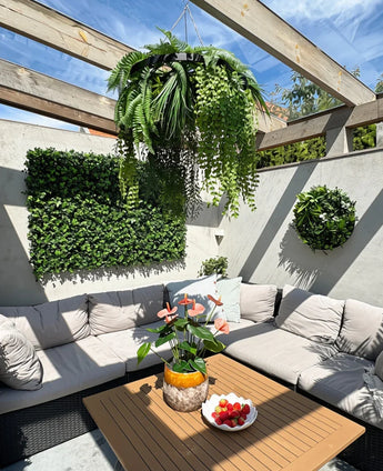 Eine sonnige Terrasse mit beigen Sitzmöbeln mit Kissen und einem Holztisch, dekoriert mit einer Ananasvase mit rosa Blumen und einer Schale mit Erdbeeren. Grüne Wandpflanzen sorgen für einen üppigen Innenraum, ergänzt durch die künstliche Hängepflanze Farn UV 60 cm im schwarzen Ring als auffälligen Hängeakzent.