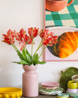 Auf einem Tisch mit buntem Geschirr steht eine rosa Vase mit der Kunstblume Französische Sittich-Tulpe 76 cm rot/gelb, während im Hintergrund teilweise ein gerahmtes Gemälde mit einem Croissant und einer Tasse zu sehen ist.