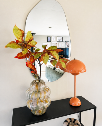 In einem gewellten Spiegel über einem schwarzen Konsolentisch befinden sich eine Glasvase mit Kunsttak Chestnut 91 cm rot/grün und eine orangefarbene Pilzlampe. Der Spiegel spiegelt ein Wohnzimmer mit gerahmter Wandkunst wider.