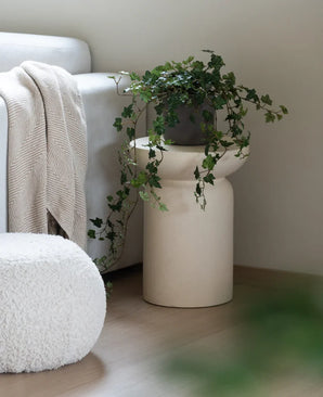 Ein künstlicher Efeu Deluxe Green 90 cm steht in einer beigen Vase auf einem Holzboden neben einem hellgrauen Sofa mit cremefarbenem Plaid und einem weißen Struktur-Pouf.