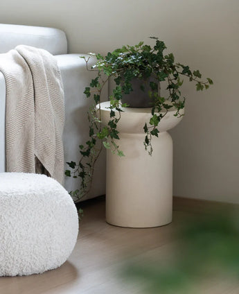 Ein künstlicher Efeu Deluxe Green 90 cm steht in einer beigen Vase auf einem Holzboden neben einem hellgrauen Sofa mit cremefarbenem Plaid und einem weißen Struktur-Pouf.