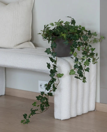 Der künstliche Efeu Deluxe Grün 90 cm mit seinen üppigen hängenden Ranken steht auf einer weiß strukturierten Bank neben einem cremefarbenen Kissen in einem minimalistischen Raum mit hellem Holzboden.