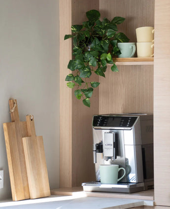 Auf der Theke steht eine silberne Kaffeemaschine mit einer grünen Tasse. Darüber sind auf einem Holzregal pastellfarbene Tassen gestapelt und eine „Art Hanging Goldranke 60 cm UV“ hängt. Direkt daneben sind hölzerne Schneidebretter an die Wand gelehnt.
