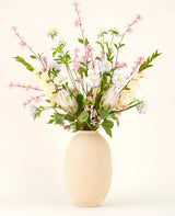 In einer Vase mit beiger Struktur befindet sich der Seiden-Kunstblumenstrauß Lots of Love XL, ein handgebundener Strauß aus rosa, weißen und blauen Seidenblumen mit grünen Blättern und langen Stielen vor hellem Hintergrund.