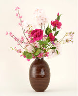 Der Kunstblumenstrauß Pink Reverie aus Seide befindet sich in einer Vase mit brauner Struktur, mit rosa, magentafarbenen und weißen Kunstblumen mit grünen Blättern vor einem schlichten hellen Hintergrund.