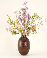 Eine braune Keramikvase zeigt den künstlichen Seidenblumenstrauß Lilac, einen hohen und eleganten Seidenstrauß mit rosa, lila und lila Blüten mit grünen Blättern und dünnen Zweigen vor einem schlichten hellen Hintergrund.