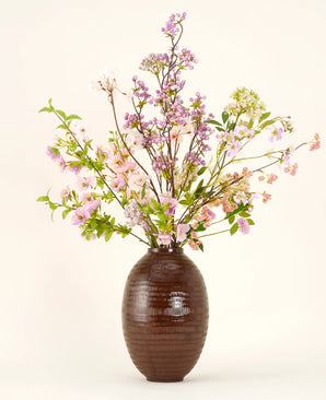Eine braune Keramikvase zeigt den künstlichen Seidenblumenstrauß Lilac, einen hohen und eleganten Seidenstrauß mit rosa, lila und lila Blüten mit grünen Blättern und dünnen Zweigen vor einem schlichten hellen Hintergrund.