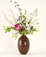 Ein künstlicher Seidenblumenstrauß von Serene Bloomfield mit einer großen rosa Protea, weißen Rosen, lila und weißen Blüten und grünem Blattwerk steht in einer braunen, gerippten Vase vor einem schlichten, hellen Hintergrund.