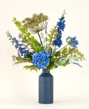 Der künstliche Blumenstrauß aus Seide von Mystic Blue Garden besteht aus blauen Hortensien, Rittersporn, grünen Blättern und getrockneten beigen Samenköpfen in einer blauen Keramikvase – ideal, um Ihren Raum vor einem hellen Hintergrund aufzuhellen.