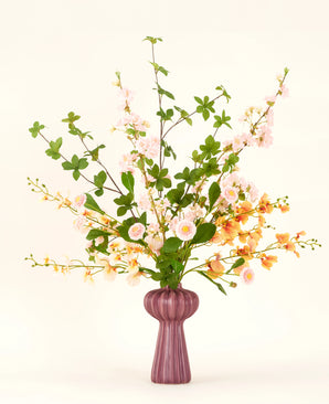 Ein Kunstblumenstrauß aus Seide von Blossom Harmony ist in einer malvenfarbenen Vase mit rosa und pfirsichfarbenen Blüten und grünen Blättern vor einem einfachen hellen Hintergrund für eine elegante Präsentation arrangiert.