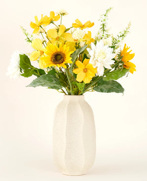 Der künstliche Blumenstrauß Sunny Whisper aus Seide mit Sonnenblumen, weißen Dahlien und grünen Blättern in einer beigen Keramikvase sticht wunderschön vor einem hellen Hintergrund hervor.