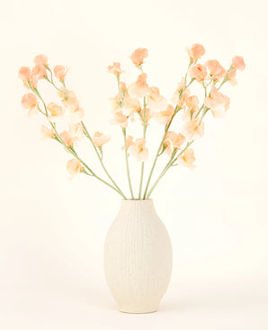 Die cremefarbene Keramikvase enthält den Kunstblumenstrauß Pea Peach aus Seide mit zarten pfirsichfarbenen und hellrosa Blüten mit langen grünen Stielen vor einem hellen, neutralen Hintergrund.