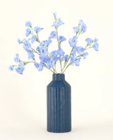 Eine blaue gerippte Keramikvase zeigt den Kunstblumenstrauß Pea Blue aus Seide mit seinen zarten hellblauen Kunstblumen vor einem schlichten, hellen Hintergrund.