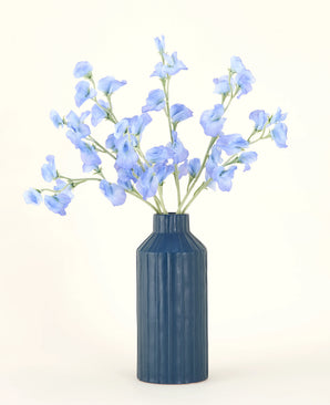 Eine blaue gerippte Keramikvase zeigt den Kunstblumenstrauß Pea Blue aus Seide mit seinen zarten hellblauen Kunstblumen vor einem schlichten, hellen Hintergrund.