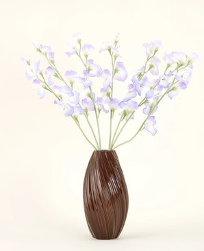 In einer braunen, gerippten Vase befindet sich der künstliche Seidenblumenstrauß Erbsen-Lavendel mit zarten Blütenblättern und grünen Stielen vor einem schlichten, cremefarbenen Hintergrund für eine dezente Landhausatmosphäre.
