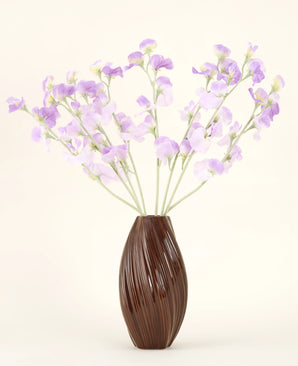 Eine strukturierte braune Vase hält den künstlichen Seidenblumenstrauß Erbsenlila-hellviolette Seidenblumen mit grünlichen Stielen vor einem schlichten, hellen Hintergrund.