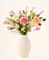 In einer weißen Keramikvase befindet sich der Strauß „Backyard Blossom“ aus künstlichen Seidenblumen, bestehend aus einem wunderschönen Arrangement aus rosa Rosen, weißen Gänseblümchen und kleinen gelben Blüten aus Seidenblumen, ergänzt durch üppige grüne Blätter vor einem schlichten hellen Hintergrund.