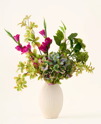 In einer weißen, strukturierten Vase steht ein Strauß grüner Hortensien, Blattzweige und leuchtend magentafarbener Seidenblumen vor einem schlichten, hellen Hintergrund. So entsteht ein auffälliger „Cottage Garden“-Strauß aus künstlichen Seidenblumen, der jedem Raum dauerhaften Charme verleiht.