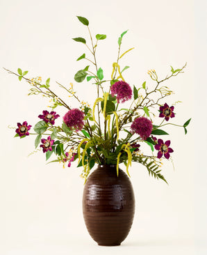 In einer braunen, gerippten Vase befindet sich der Strauß „Woodland Charm“ aus künstlichen Seidenblumen mit lila Allium, dunkelrosa Blüten, gelben Zweigen und grünen Blättern vor einem schlichten, hellen Hintergrund.