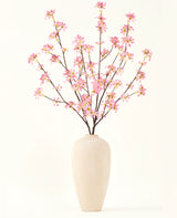 Eine hohe beige Keramikvase enthält mehrere Zweige des künstlichen Blumenstraußes aus Seide. Rosa Blumen mit zartrosa Blüten und grünen Blättern vor einem schlichten weißen Hintergrund.
