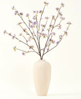 Eine hohe, cremefarbene Keramikvase enthält eine Reihe dünner Zweige mit dem Text „Seiden-Kunstblumenstrauß“ Künstlicher Blumenzweig lila, vor einem schlichten, hellen Hintergrund.