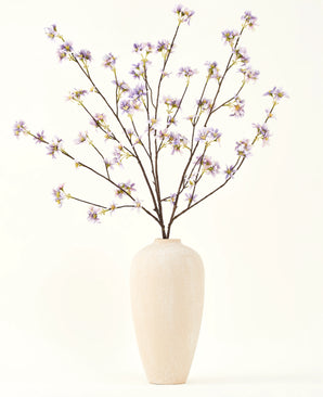 Eine hohe, cremefarbene Keramikvase enthält eine Reihe dünner Zweige mit dem Text „Seiden-Kunstblumenstrauß“ Künstlicher Blumenzweig lila, vor einem schlichten, hellen Hintergrund.