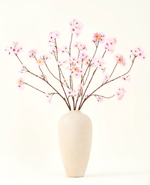 Der Seiden-Kunstblumenstrauß Kirschblütenrosa 114 cm hat lange, dünne Zweige mit Büscheln hellrosa Blüten in einer beigen Keramikvase, die einen auffälligen Kontrast zum schlichten weißen Hintergrund bilden.