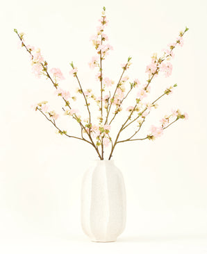 Der Seidenblumenstrauß Kirschblüten hellrosa 94 cm besteht aus zartrosa künstlichen Kirschblüten in einer weißen Keramikvase, die vor einem schlichten hellen Hintergrund Eleganz verleiht.