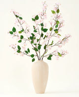 Eine beige Keramikvase enthält mehrere lange Zweige mit dem künstlichen Seidenblumenstrauß aus Pfirsichblüten-Rosa und grünen Blättern vor einem schlichten weißen Hintergrund – perfekt für ein Interieur mit künstlichen Blumen.