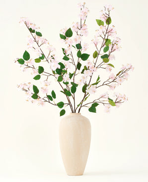 Eine beige Keramikvase enthält mehrere lange Zweige mit dem künstlichen Seidenblumenstrauß aus Pfirsichblüten-Rosa und grünen Blättern vor einem schlichten weißen Hintergrund – perfekt für ein Interieur mit künstlichen Blumen.