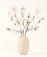 Der Seiden-Kunstblumenstrauß Cherry Blossom Blush 119 cm zeigt zarte, hellrosa Kirschblüten mit grünen Blättern in einer beigen Vase, ideal als künstliche Blumendekoration vor hellem Hintergrund.