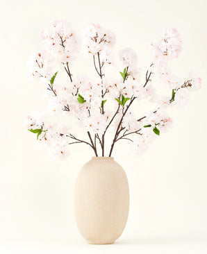 Der Seiden-Kunstblumenstrauß Cherry Blossom Blush 119 cm zeigt zarte, hellrosa Kirschblüten mit grünen Blättern in einer beigen Vase, ideal als künstliche Blumendekoration vor hellem Hintergrund.