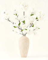 Der künstliche Seidenblumenstrauß Kirschblütencreme 119 cm besteht aus eleganten braunen Zweigen mit zartweißen Kunstblüten und grünen Blättern, die in einer schlichten beigen Vase vor einem schlichten hellen Hintergrund präsentiert werden.