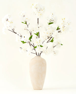 Der künstliche Seidenblumenstrauß Kirschblütencreme 119 cm besteht aus eleganten braunen Zweigen mit zartweißen Kunstblüten und grünen Blättern, die in einer schlichten beigen Vase vor einem schlichten hellen Hintergrund präsentiert werden.