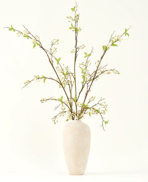 In einer hohen, cremefarbenen Keramikvase befindet sich der künstliche Seidenblumenstrauß Verbena Creme mit langen Zweigen, kleinen grünen Blättern und zarten weißen Blüten, wunderschön arrangiert vor einem schlichten hellen Hintergrund.