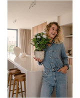 Eine lächelnde Frau in einem Jeansoverall steht in einer modernen Küche, mit der weißen Kunstpflanze Artificial Gardenia UV 39 cm in ihren Händen. Auf einer weißen Arbeitsplatte sind Holzhocker aufgereiht, durch ein Fenster im Hintergrund strömt Sonnenlicht herein.