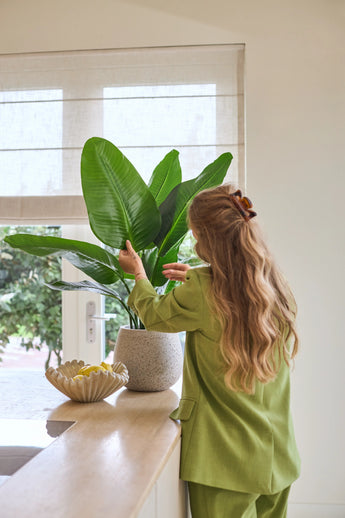 Eine Frau mit langen, welligen Haaren, gekleidet in einen grünen Anzug, kümmert sich um eine 90 cm große Strelitzia Real Touch Deluxe-Kunstpflanze auf einer Theke neben einer Schale mit Zitronen in einem hellen Raum mit Fenster.