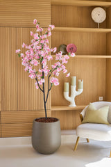 Eine künstliche Kirschblüte 180 cm rosa, ein Topfbaum mit rosa Blüten, steht neben einem weißen Stuhl mit Kissen in einem modernen Raum mit Holzregalen und Dekogegenständen.