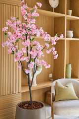 Die künstliche Kirschblüte 150 cm rosa sieht mit ihren zartrosa Seidenblüten elegant in einem modernen Raum mit Holzregalen, Dekorationsgegenständen, einem beigen Stuhl und einem grünen Kissen aus und verleiht Ihrem Interieur den Charme von Seidenblumen.