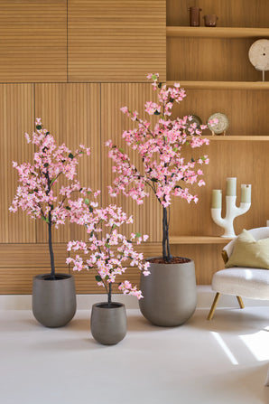 Drei Topfpflanzen vom Typ Kunstpflanze Bougainvillea, 120 cm groß, rosafarben und mit rosa Blüten, stehen auf einem weißen Boden in einem modernen Raum mit Holzvertäfelung, Regalen und einem weißen Stuhl.