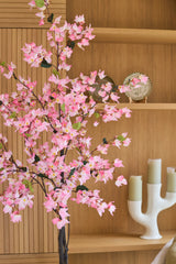 Eine 150 cm große künstliche Bougainvillea-Pflanze in rosa Farbe mit rosa Blüten steht vor Holzregalen, auf denen ein dekorativer Teller und ein moderner weißer Kerzenständer mit drei grünen Kerzen ausgestellt sind.