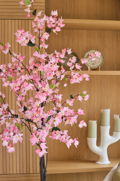 Eine 150 cm große künstliche Bougainvillea-Pflanze in rosa Farbe mit rosa Blüten steht vor Holzregalen, auf denen ein dekorativer Teller und ein moderner weißer Kerzenständer mit drei grünen Kerzen ausgestellt sind.